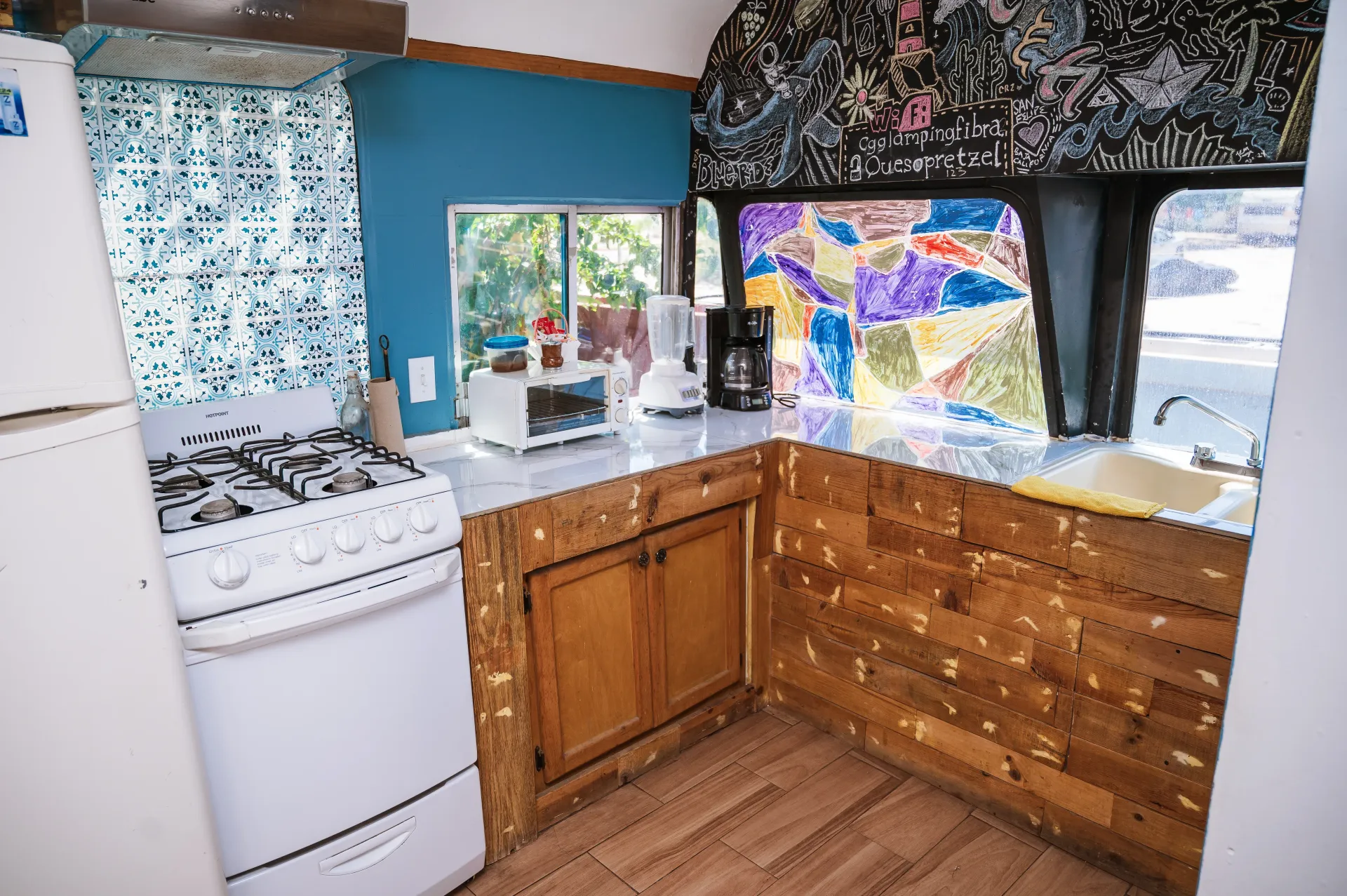 Small kitchen inside a bus with stove, cabinets, and counter. Window has a mosaic-like design.