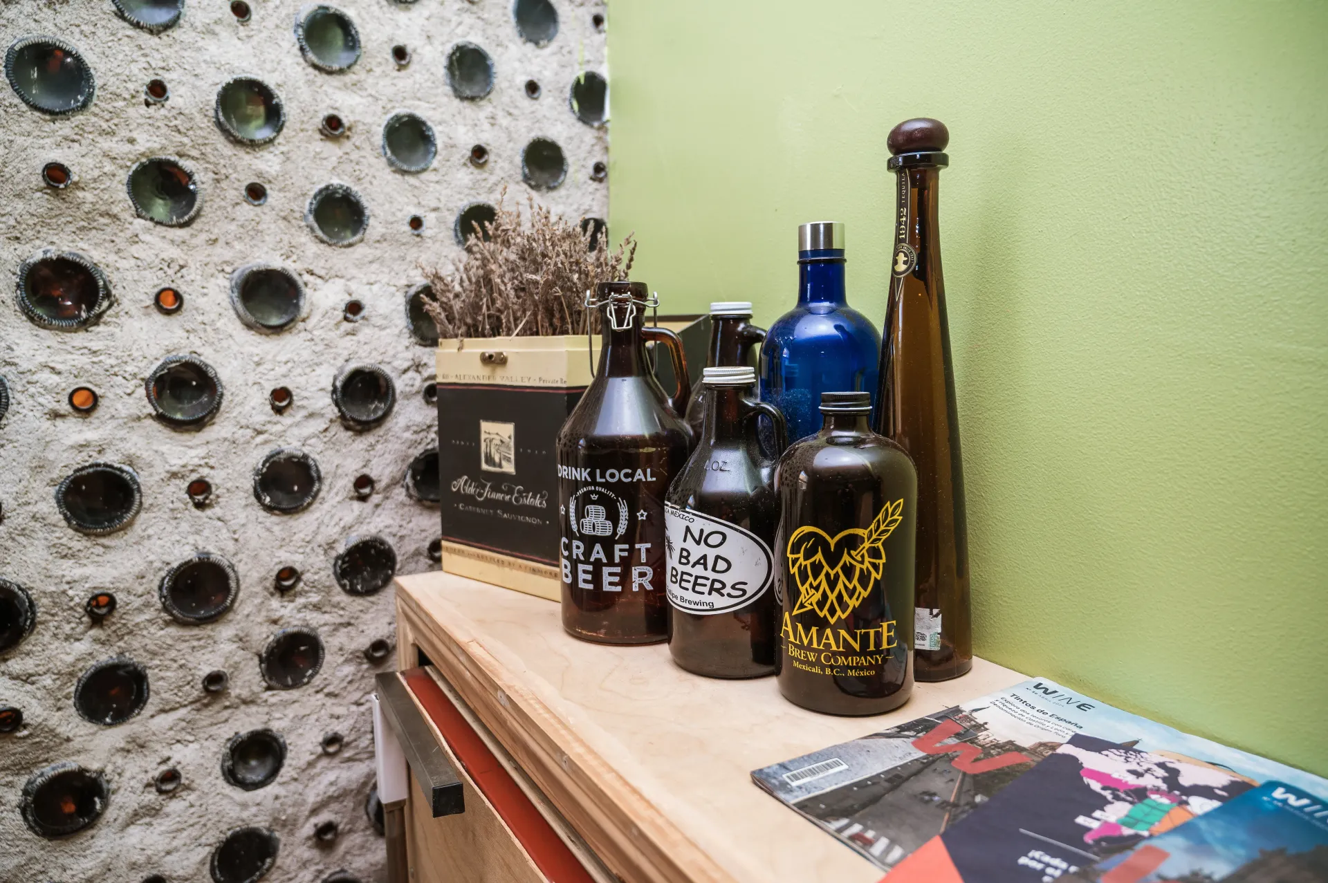 Bottles of alcohol on a wooden shelf against a wall with glass bottle embedded decor.