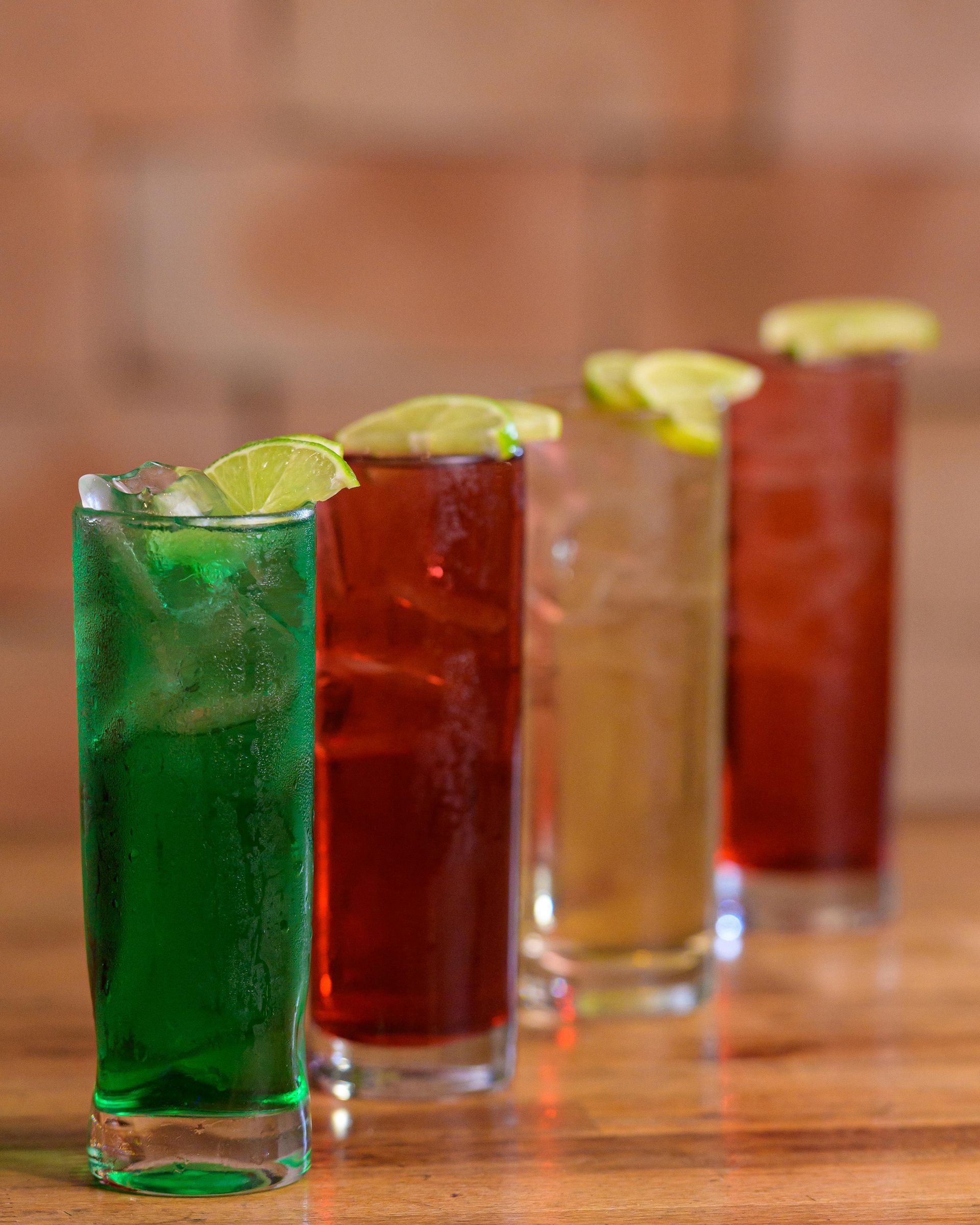 Four colorful cocktails in tall glasses, each with a lime garnish, lined up on a wooden surface.