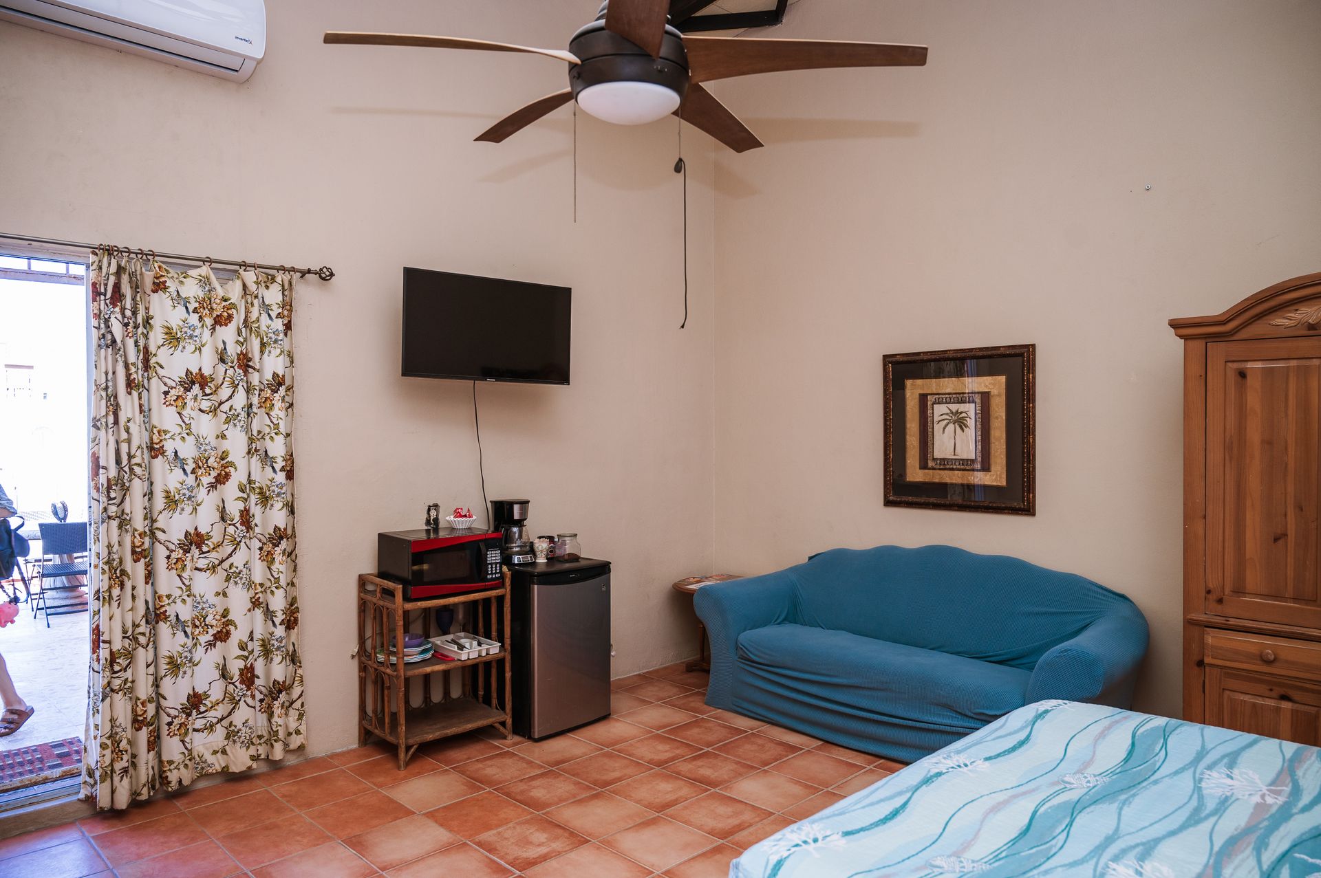 Bedroom with blue sofa, TV, mini-fridge, open door to patio, and ceiling fan.