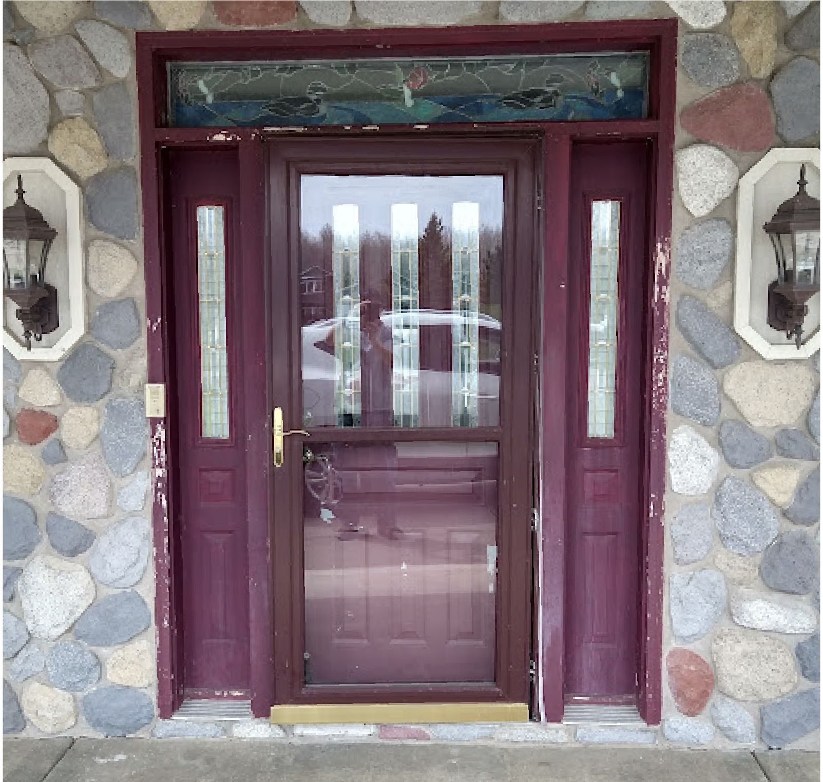 A dark red front door with glass panels and a screen door, set in a stone exterior wall with wall-mounted lanterns.