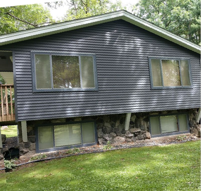 A split-level house with dark grey horizontal siding, stone foundation, and a wooden deck, set against a green lawn.