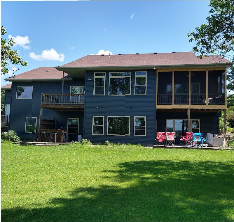 A two-story dark blue house with a wooden deck and a screened-in porch, overlooking a green lawn under a sunny sky.