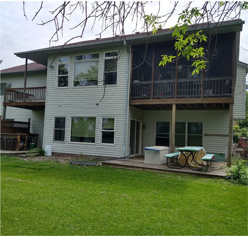 A two-story house with off-white siding, a screened-in porch, a wooden deck, and a patio with a picnic table in a backyard.