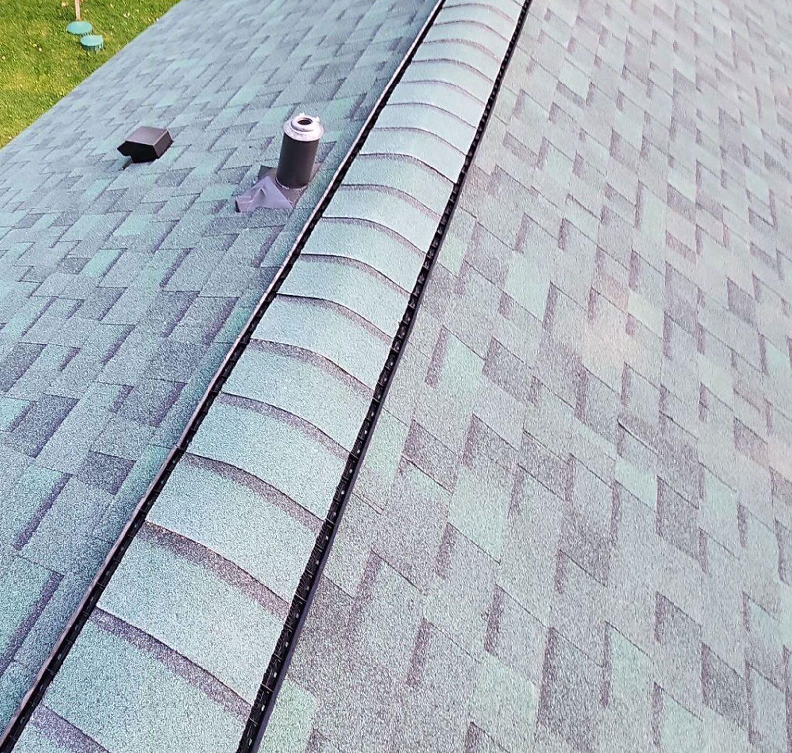 A view from above showing a grey shingled roof with a central ridge vent, a plumbing stack, and a vent cover.