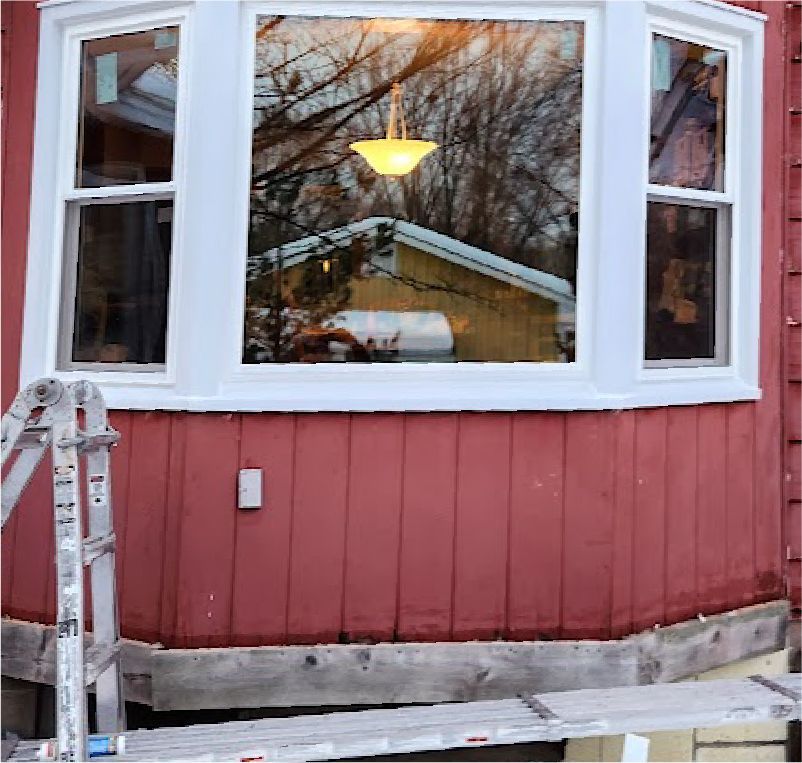 A bay window on a red-sided house with a white frame, under renovation with scaffolding and a ladder in the foreground.