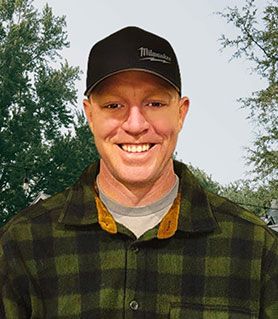 A smiling person wearing a black Milwaukee-branded baseball cap and a green and black plaid shirt against a tree background.