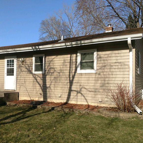 A tan-sided house exterior with a white door, two windows, a brown roof, and a downspout, seen on a sunny day.