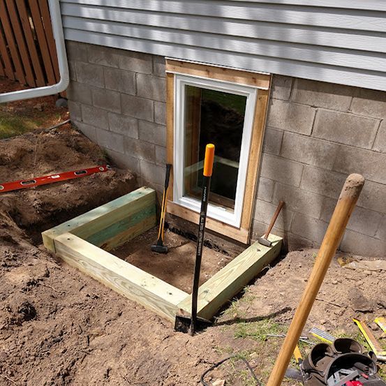 A window well under construction with pressure-treated lumber framing outside a basement window in a concrete block wall.
