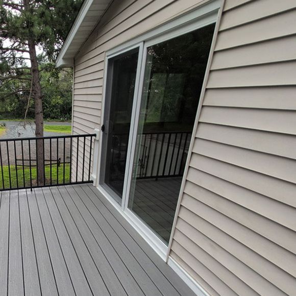 A gray composite deck with a black railing, featuring a white sliding glass door set into light tan vinyl siding.