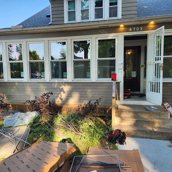 A front view of a house with gray siding, multiple windows, an open door, and construction materials on the front lawn.