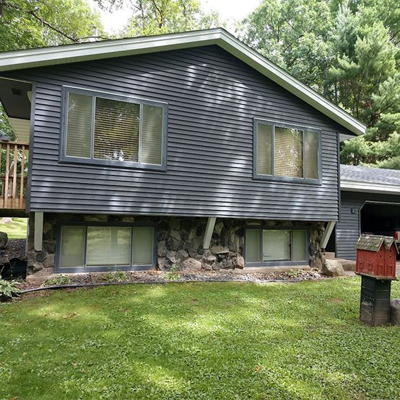 A two-story house with dark grey horizontal siding, stone foundation, and multiple windows set in a green lawn.