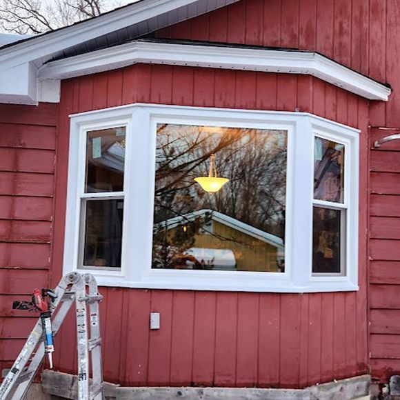A new white bay window installed on a red wooden house exterior, with a ladder and caulk gun nearby.