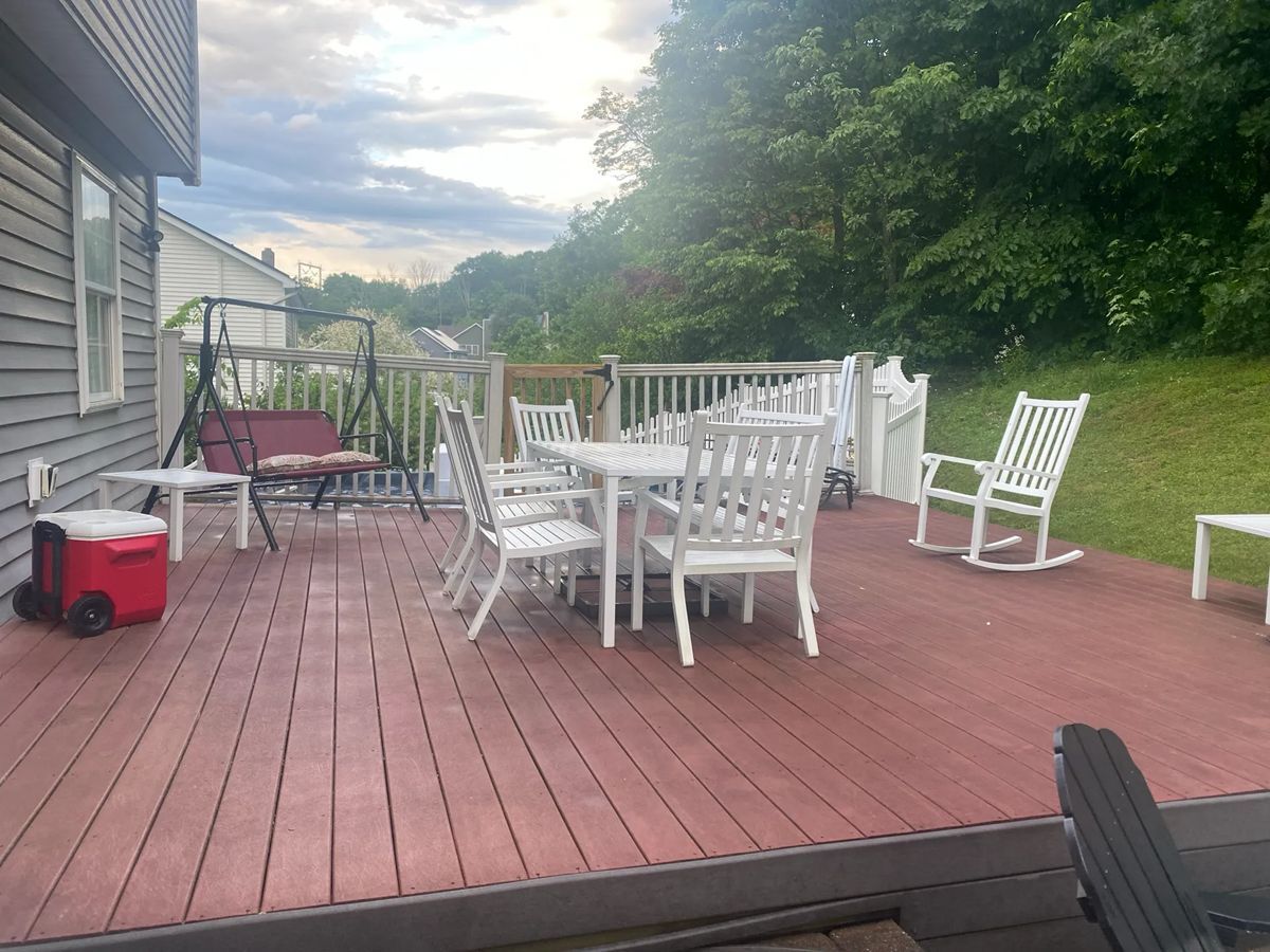 Wooden deck with white furniture, a red cooler, and a swing. A grassy hill and trees are in the background.