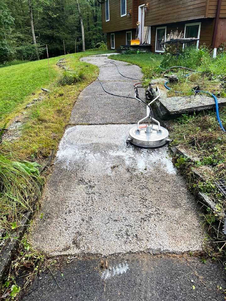 A person power washing a concrete walkway next to a grassy lawn and house with a deck.