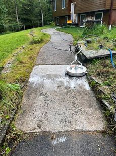 A person power washing a concrete walkway next to a grassy lawn and house with a deck.