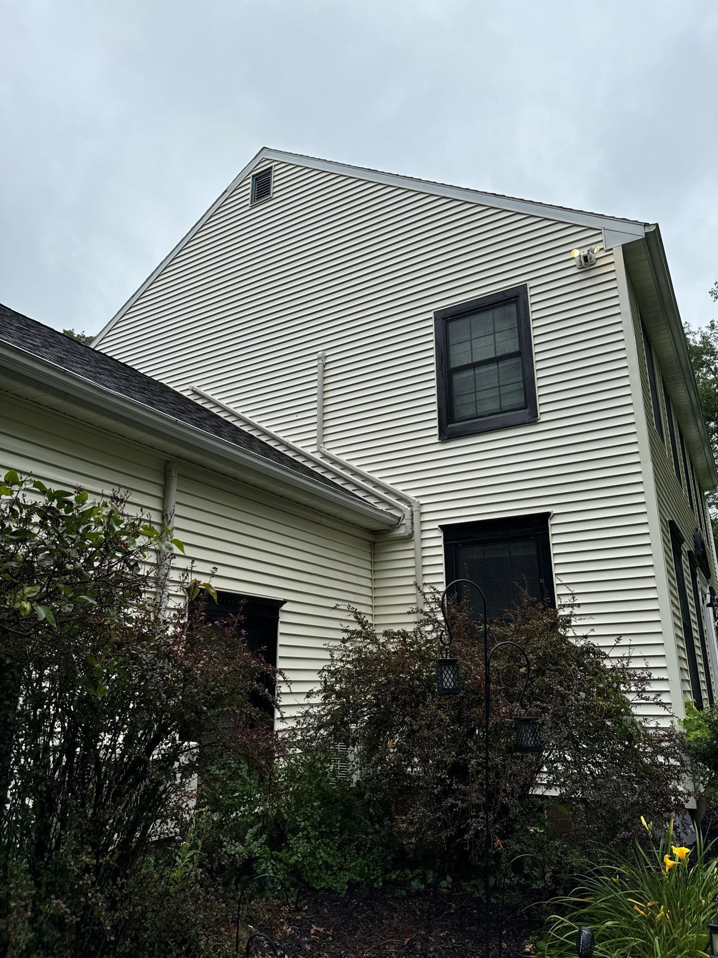Two-story house with light siding and black window frames against a cloudy sky, surrounded by bushes.