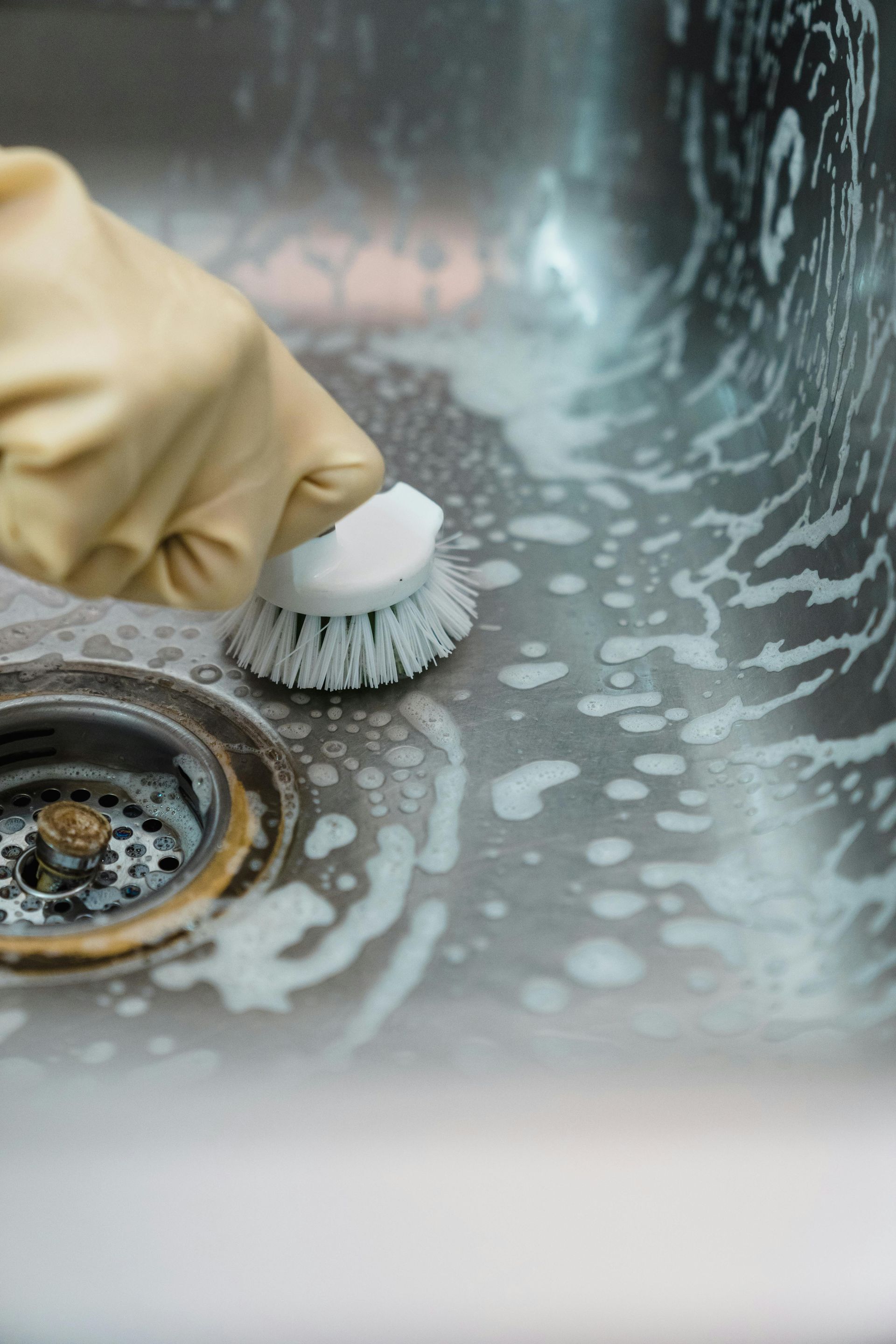 Person in glove scrubbing a stainless steel sink with a brush, soapy water visible.