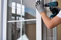 Person in gloves uses a drill to install a white window frame.