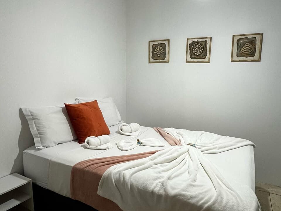 Uma cama impecavelmente arrumada com lençóis brancos, um travesseiro cor de ferrugem e toalhas dobradas, com três gravuras fixadas na parede.