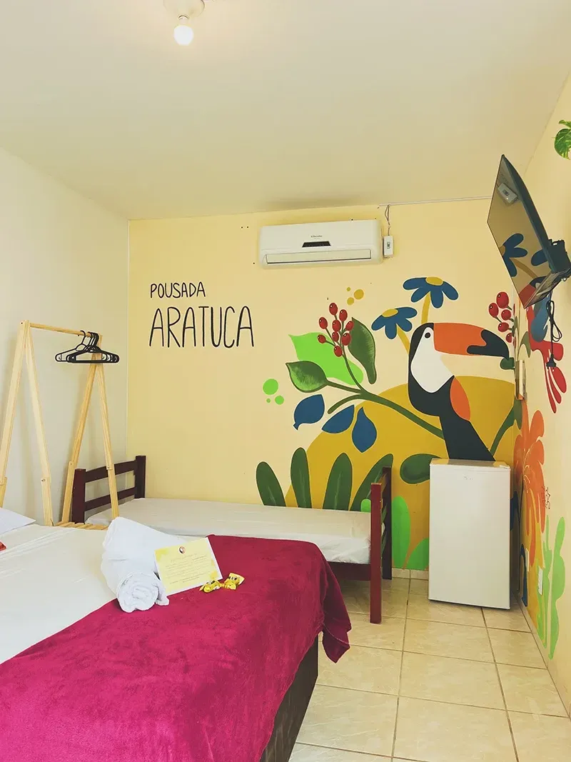 Um quarto de hotel com um mural de um tucano na parede, apresentando uma cama grande com um cobertor rosa, uma cama de solteiro e uma TV.