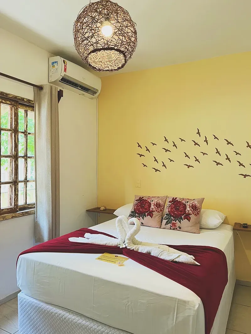 Um quarto de hotel com uma cama queen-size, um caminho de mesa vermelho, almofadas florais e cisnes de toalha contra uma parede amarela com adesivos de pássaros.