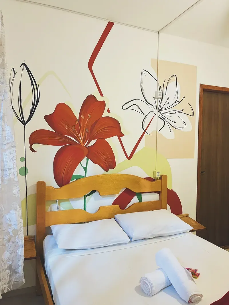 Uma cama com lençóis brancos e duas toalhas enroladas em frente a uma parede pintada com um grande lírio vermelho e desenhos florais.