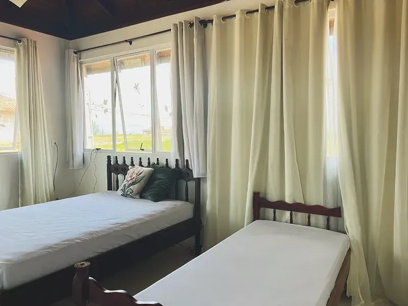 Um quarto com duas camas de estrutura de madeira, uma delas com travesseiros, e grandes janelas cobertas por cortinas cor creme.