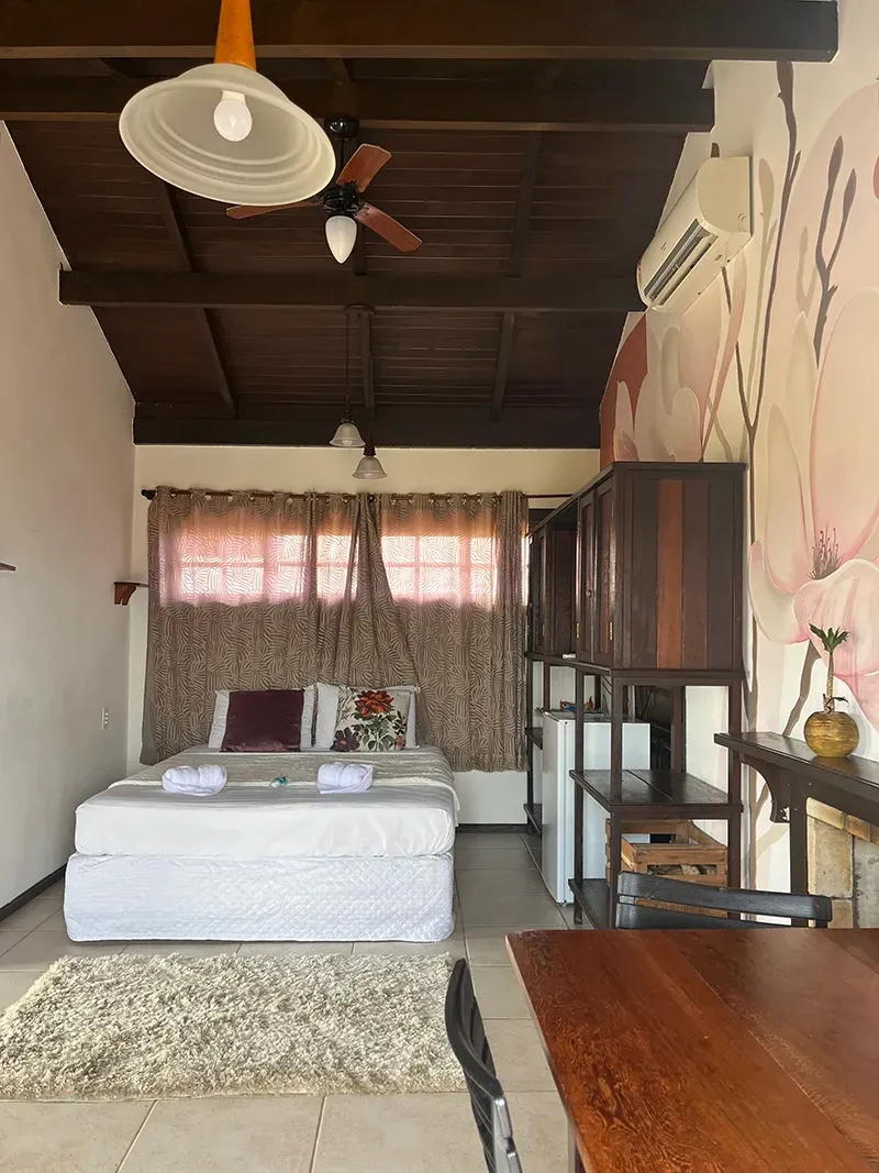 Um quarto com roupa de cama branca, um tapete estampado, vigas de madeira escura no teto, um ventilador de teto e um mural na parede lateral.