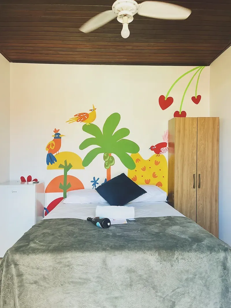Um quarto com cama de casal central, guarda-roupa de madeira, armário branco e um mural colorido com tema da natureza na parede.