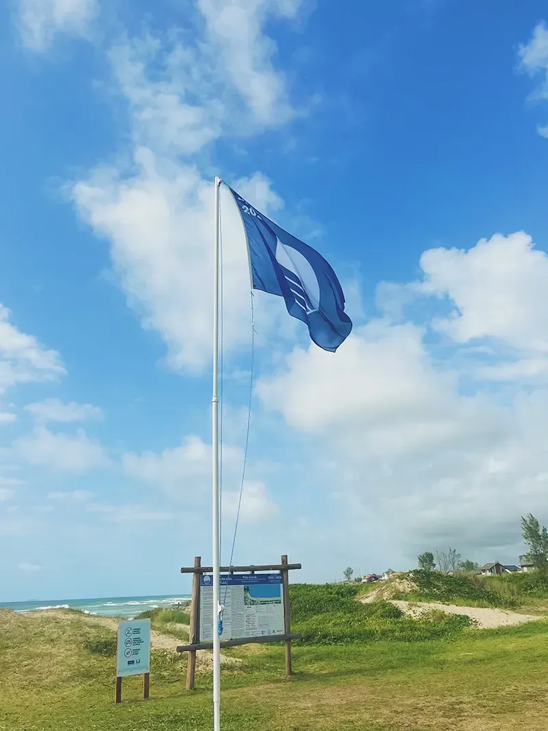 Uma bandeira azul hasteada em um mastro em uma praia costeira com dunas e um céu ensolarado e nublado ao fundo.
