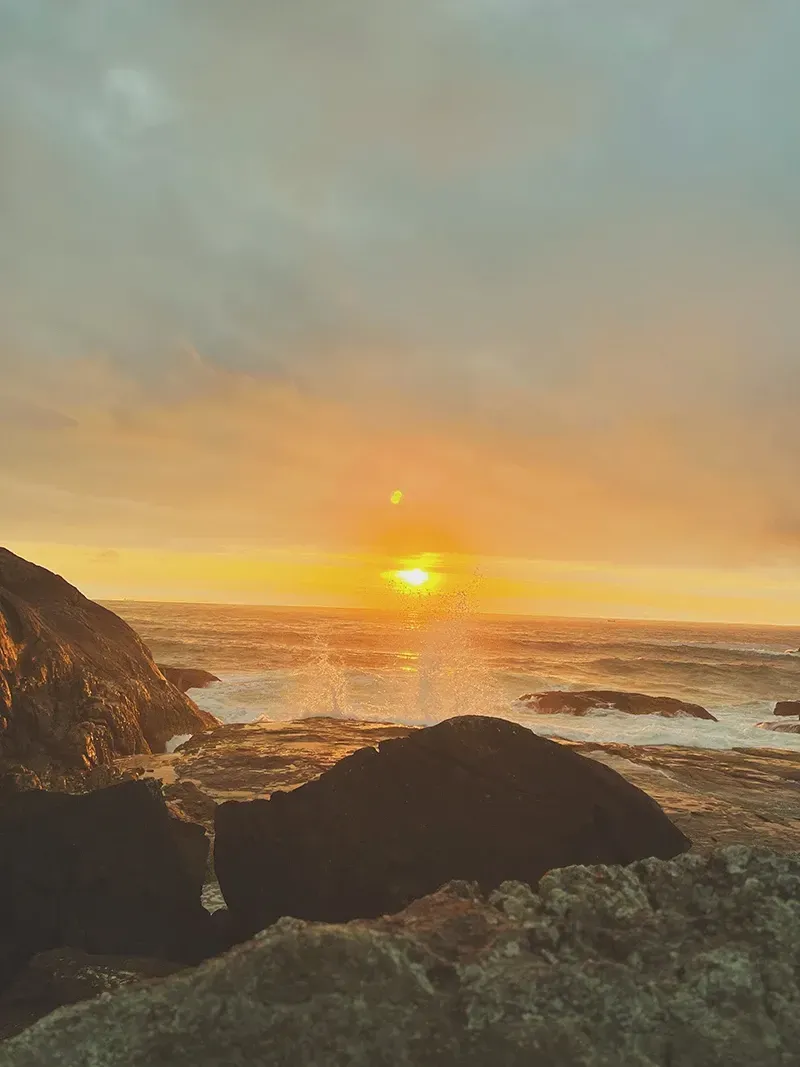 Um pôr do sol dourado ilumina uma costa rochosa, com ondas quebrando contra as silhuetas escuras das rochas litorâneas.