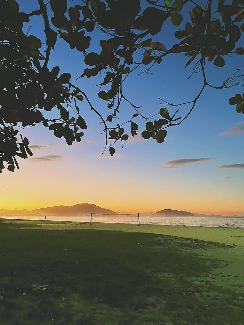 Um pôr do sol sereno sobre uma margem gramada, com galhos de árvores emoldurando o céu e montanhas distantes no horizonte.