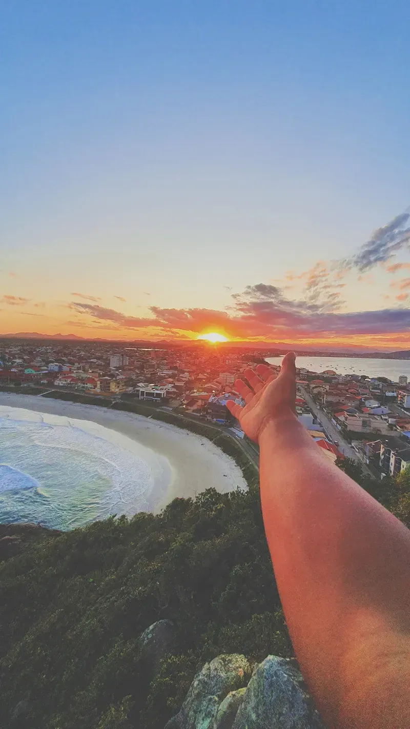 O braço de uma pessoa estende-se em direção a um pôr do sol vibrante sobre uma cidade costeira e sua praia, visto de um ponto de vista elevado.