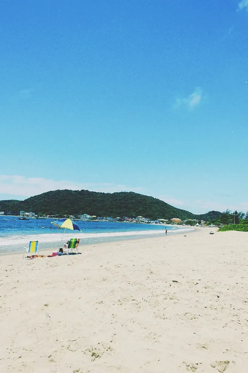 Uma praia ensolarada com areia clara, mar azul e uma colina verde ao longe sob um céu azul límpido, com um guarda-sol colorido.