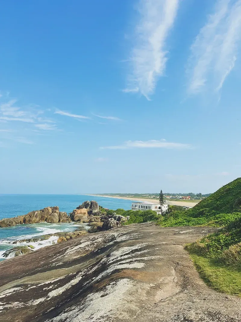 Um penhasco rochoso com vista para um oceano azul com ondas quebrando na costa, um prédio ao longe e um céu claro e nublado.