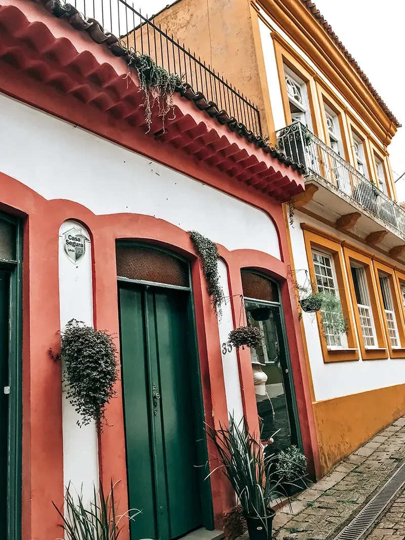 Edifícios em estilo colonial com paredes brancas, vermelhas e amarelas, portas verdes e varandas de ferro forjado em uma rua de pedra.