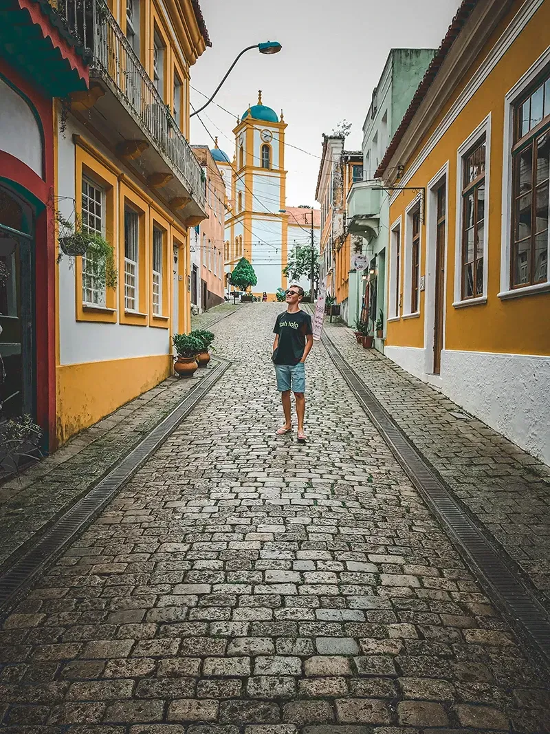 Uma pessoa está no centro de uma rua histórica de paralelepípedos, ladeada por edifícios coloridos em estilo colonial.