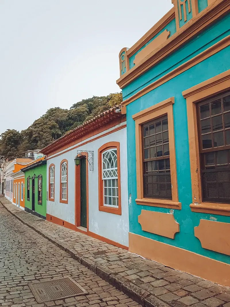 Uma rua com edifícios coloridos em estilo colonial em tons de verde-azulado, azul-claro e verde, com detalhes em madeira escura e caminhos de paralelepípedos.