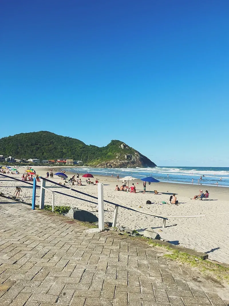 Um dia ensolarado em uma praia de areia com pessoas sob guarda-sóis, que leva às ondas e a uma colina verde sob um céu azul claro.