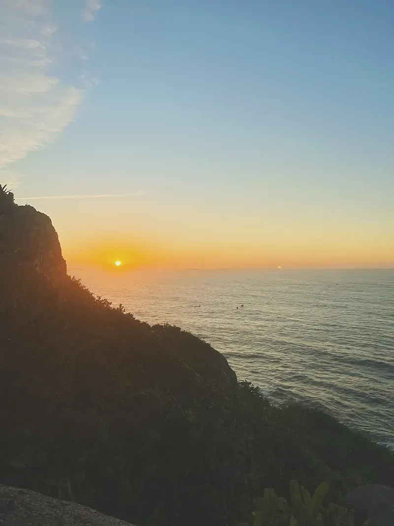 Um pôr do sol resplandece no horizonte, lançando uma luz dourada e quente sobre o oceano ao lado de um penhasco costeiro escuro e rochoso.
