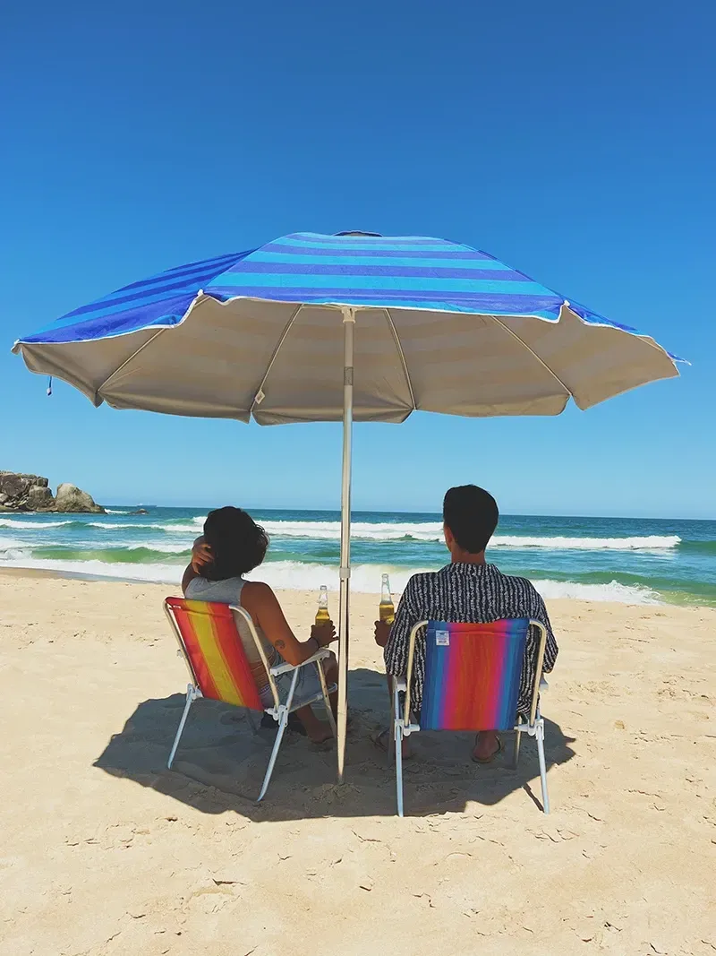 Duas pessoas sentam-se em cadeiras de praia sob um guarda-sol listrado em azul e branco, segurando bebidas enquanto contemplam o oceano.