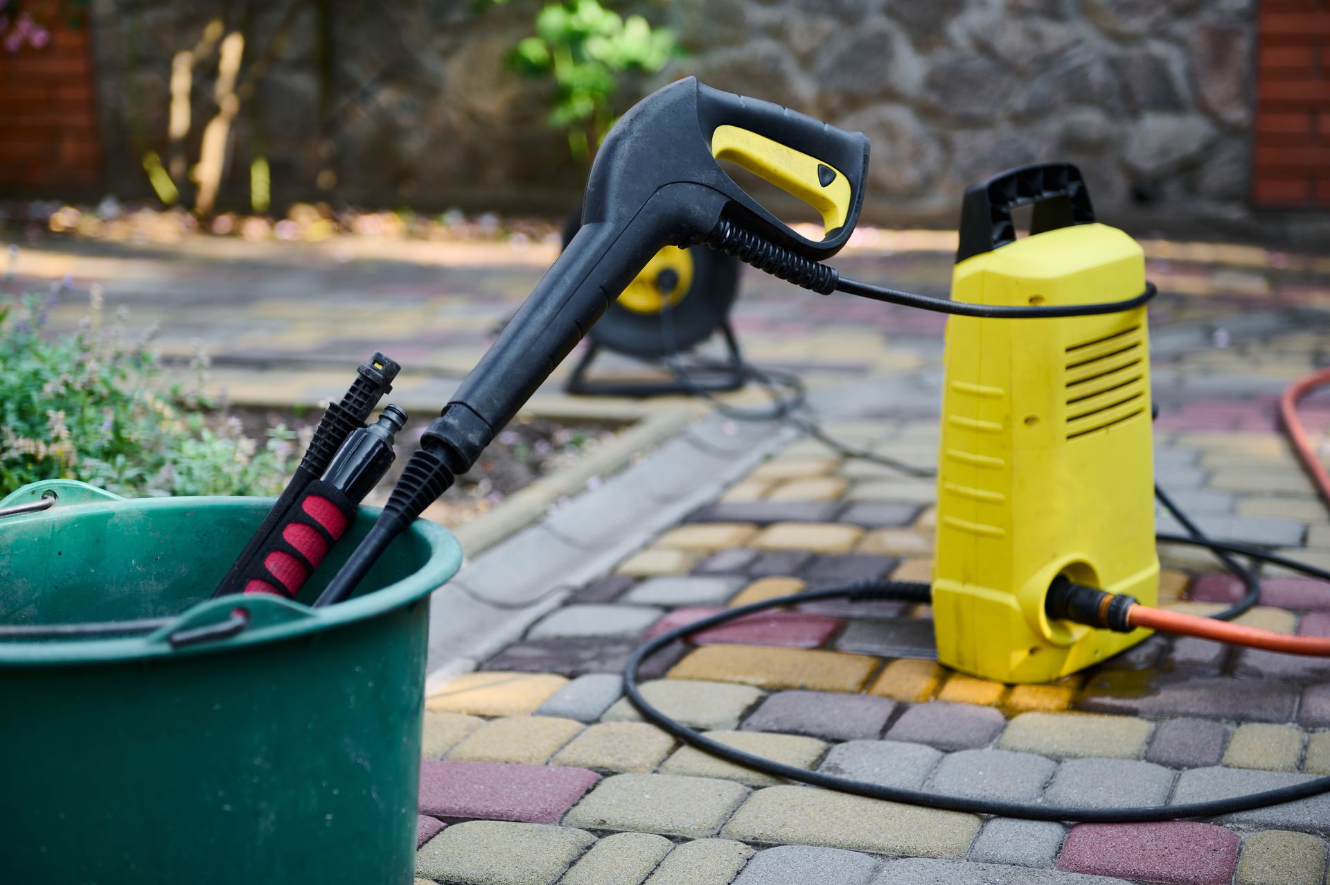 Yellow pressure washer on a stone patio spraying water into a green bucket with garden tools.