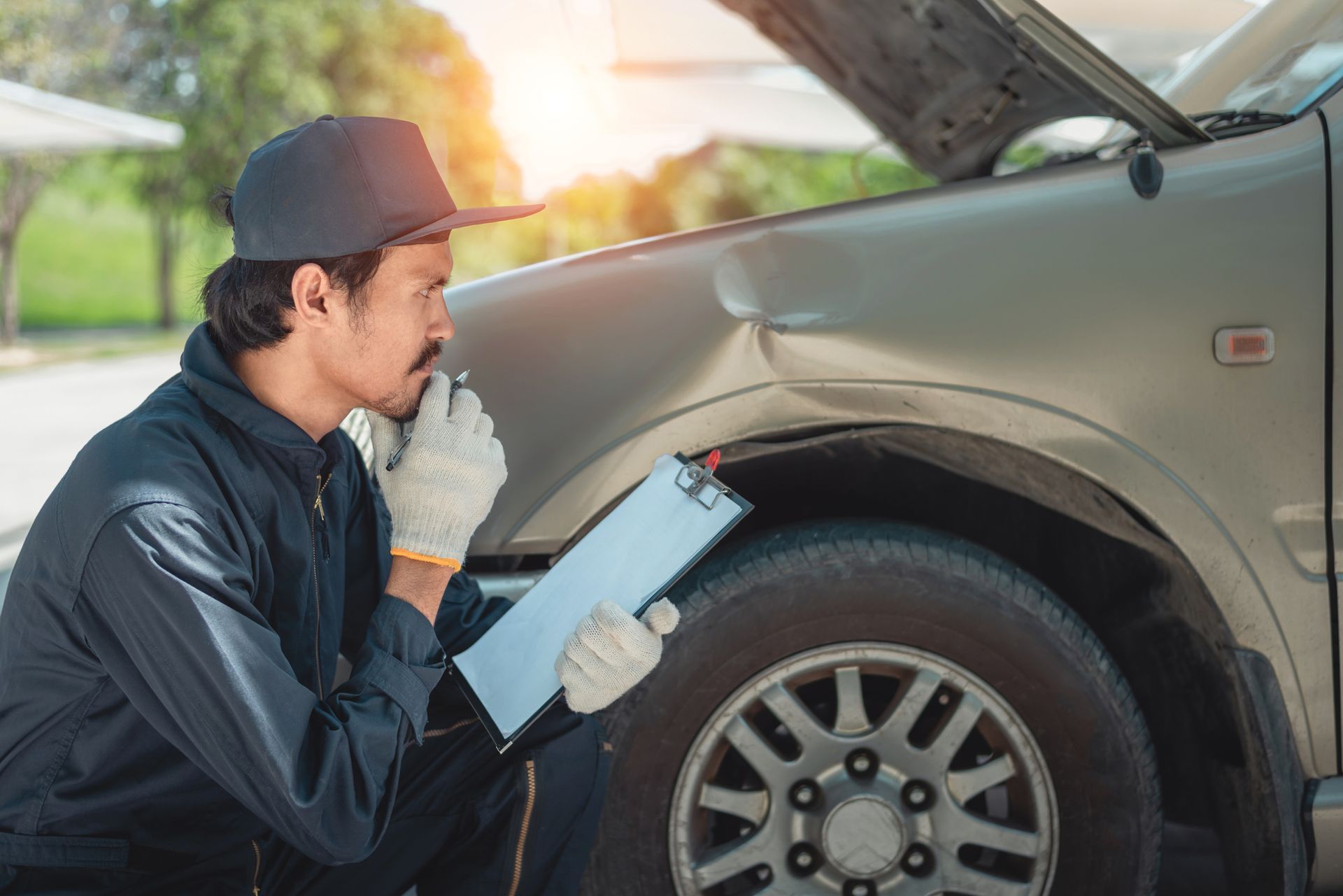 A mechanic inspecting a dented car fender with a clipboard, hood open, working outdoors on damage.