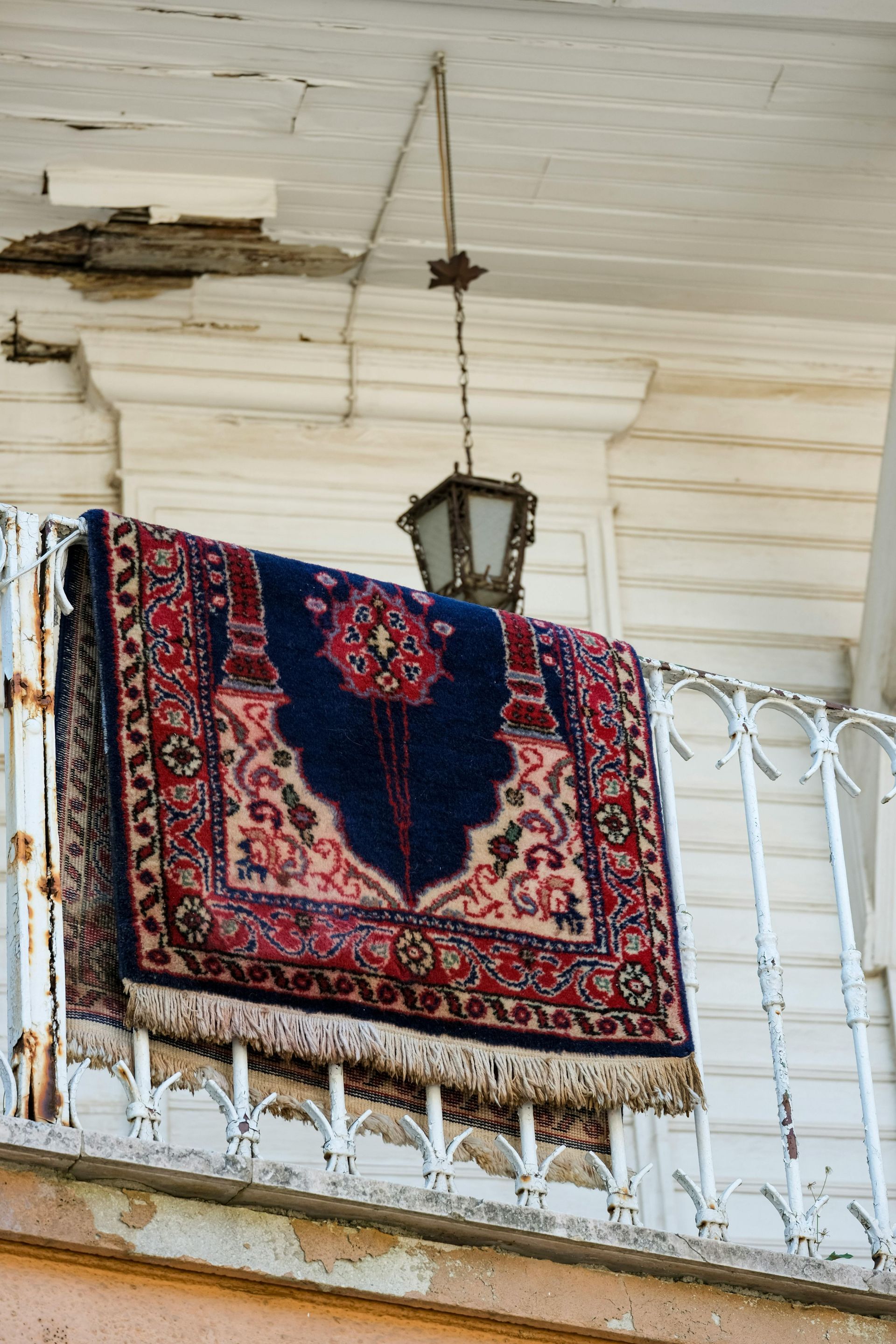 Patterned rug draped over a white balcony railing beneath a lantern on a weathered porch ceiling