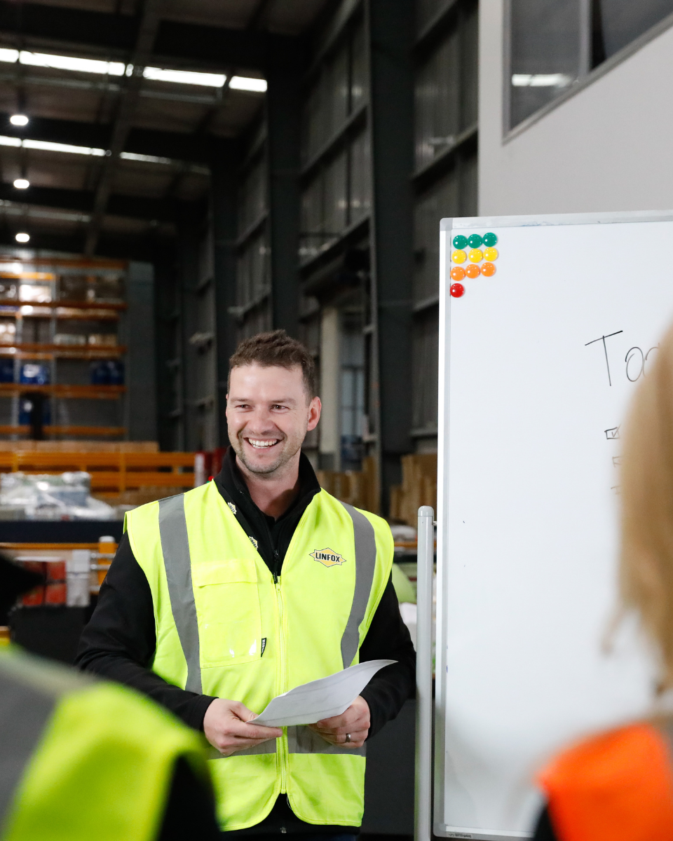 A man in a yellow vest is smiling in front of a white board