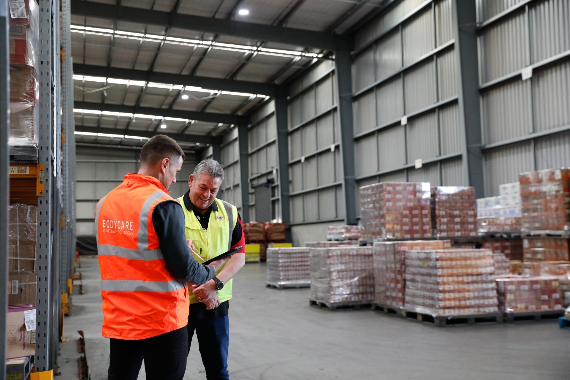 Two men are standing in a warehouse looking at a tablet. Bodycare and the client.