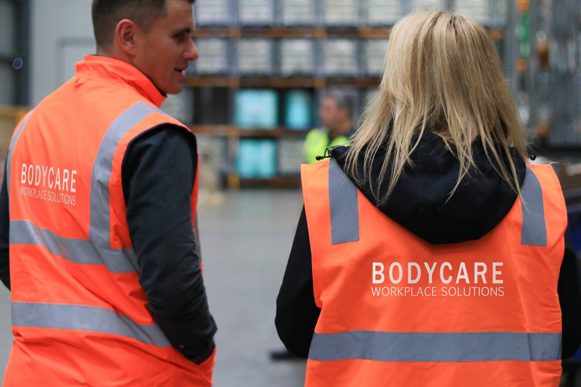 A man and a woman wearing bodycare safety vests are standing next to each other in a warehouse.