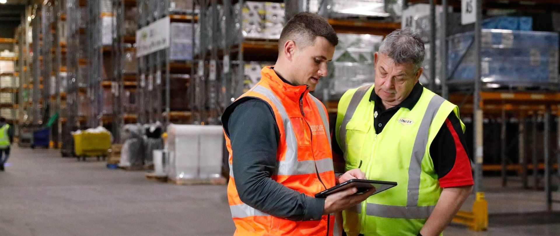 Two men are standing in a warehouse looking at a tablet. Bodycare and the client.