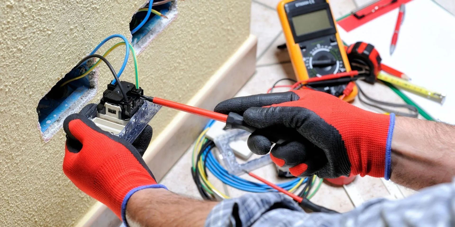 Electrician working on wiring a wall outlet, wearing red gloves.